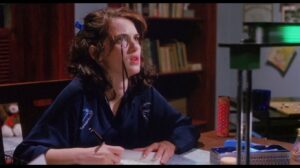 Actress Winona Ryder, portraying Veronica Sawyer in the film Heathers, sits at a desk and writes in a diary. She has long brown hair and is wearing a blue robe and a monocle.